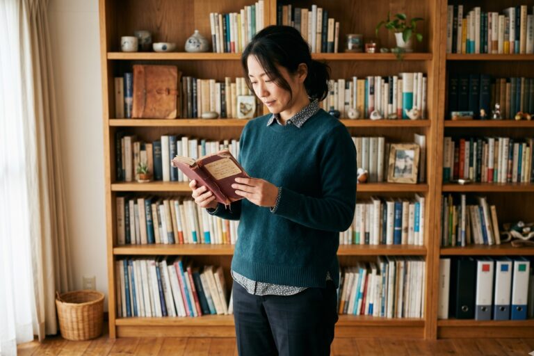 本棚の前で古い本を手に取り、手放すか迷うように見入る女性。保有効果（手放せないモノへの愛着）を表すイメージ