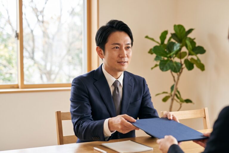 打ち合わせの席で、テーブル越しにクライアントへ資料のフォルダを手渡す日本人のビジネスパーソン
