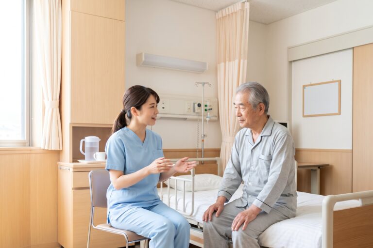 病室で高齢の男性患者の隣に座り、やさしく説明する看護師
